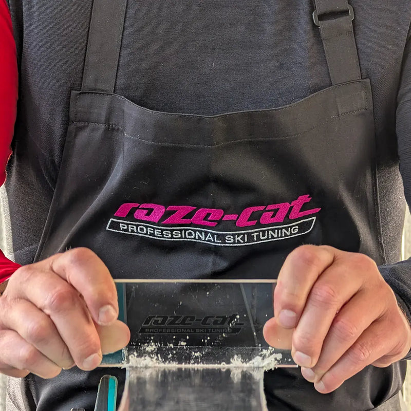 Chargez l'image dans le visualiseur de galerie, Skiservice Mensch trägt schwarze Schürze mit aufgesticktem raze-cat Logo auf der Brust (magenta und weiß). Er schabt überschüssiges Wachs vom Ski mit einer Wachsklinge (Stück Plexiglas).