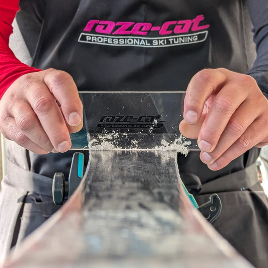 Wachsklinge (Plexiglas) wird über gewachsten Ski geschoben. Wachsspäne enstehen an der Kante. Serviceperson trägt schwarze Schürze mit magenta raze-cat Logo. 